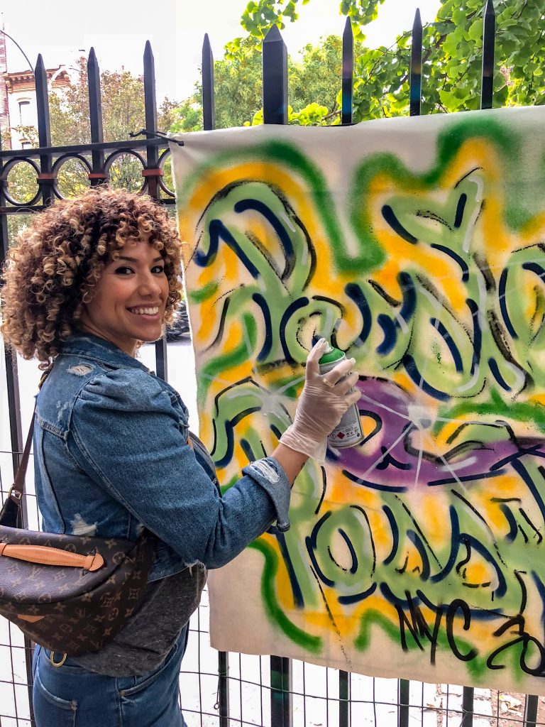 Woman wearing Louis Vuitton bum bag, while spray painting graffiti on a canvas affixed to a fence in a New York City park.