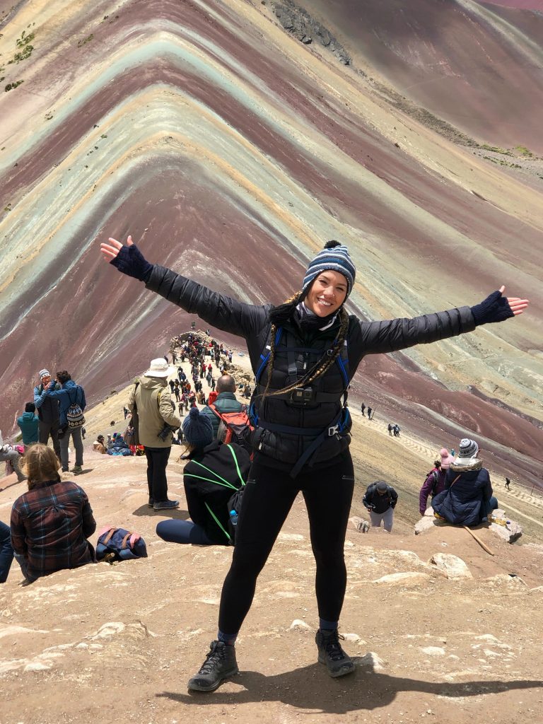 A woman who hiked to top of Rainbow Mountain with arms stretched in the air with a sense of accomplishment wearing a Go Pro chest mount.